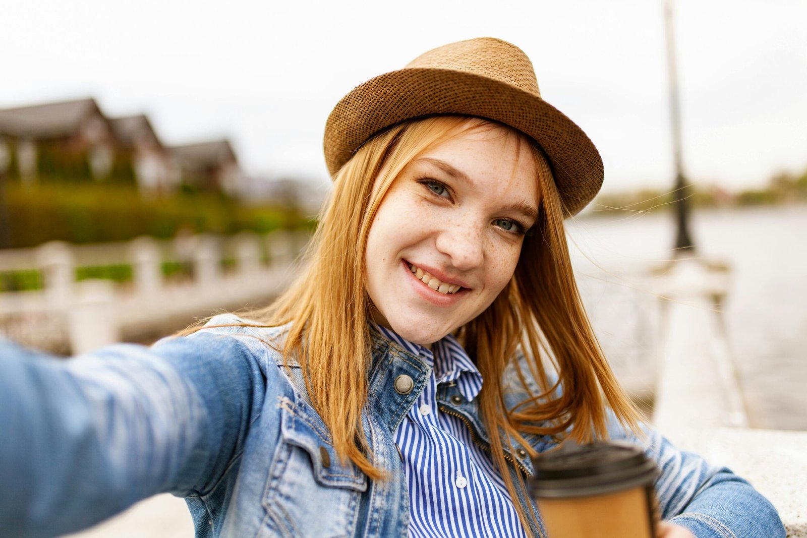 Young woman in a fedora hat takes a selfie with a coffee by the lake.